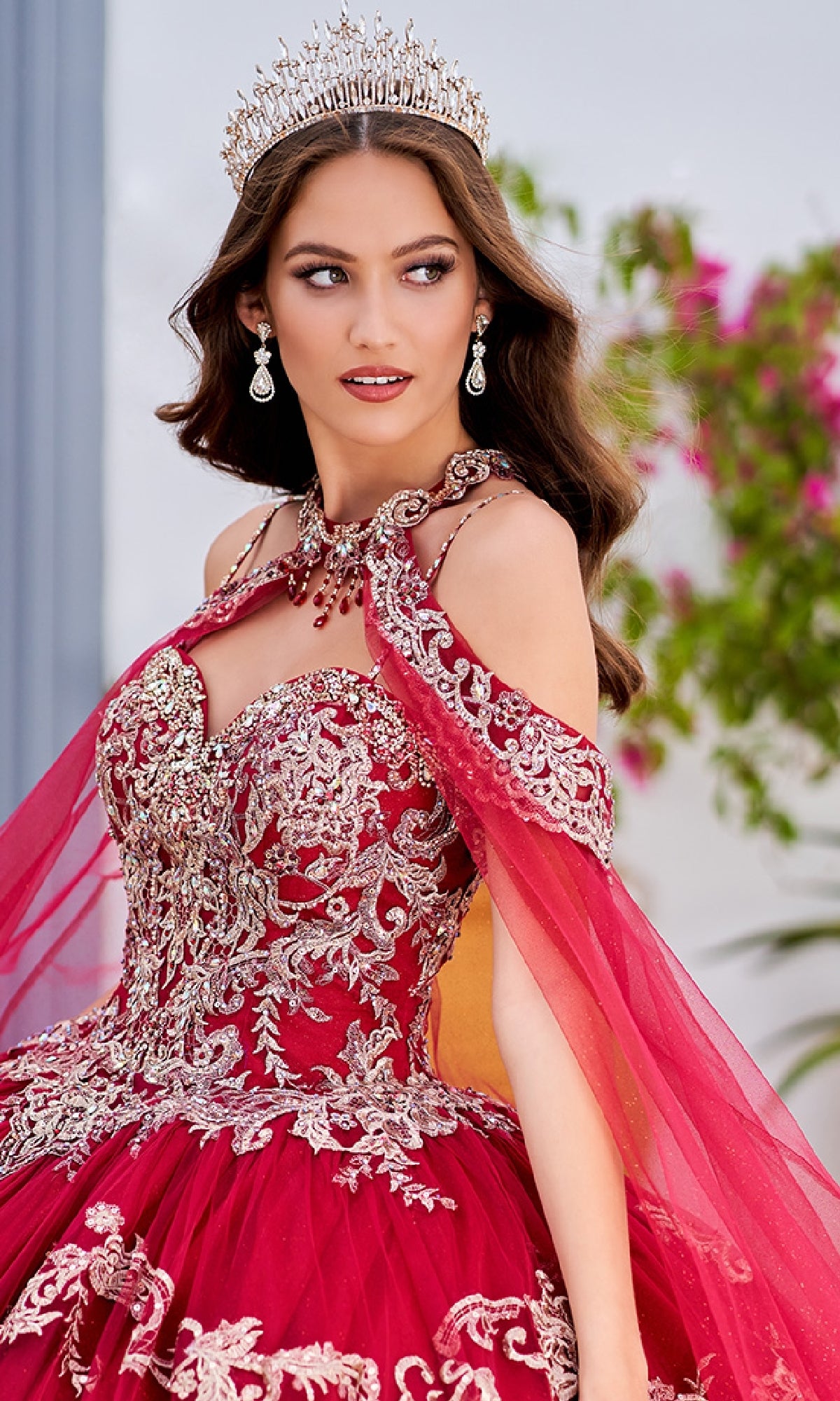 A woman stands outdoors near greenery wearing the Quinceanera Dress PR30086 By Princesa in ornate red and gold with embroidered lace, a matching sheer cape, jeweled necklace, earrings, and a crown.